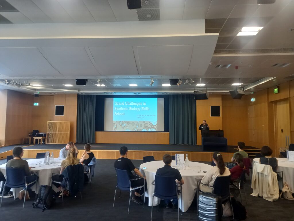 Room filled with round tables and people sat at them facing projector screen. Projector screen displays the words Grand Challenges in Synthetic Biology skills school.